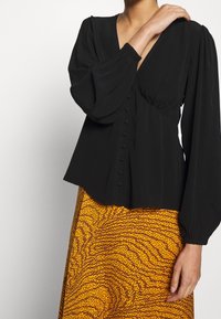 Blouse noire à col en V, manches longues bouffantes et détails de boutons sur le devant, associée à une jupe jaune à motif à pois.