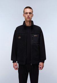 Black fleece vest with a high collar, textured surface, front zipper, and logo patch. Worn over a black long-sleeve shirt and black pants.