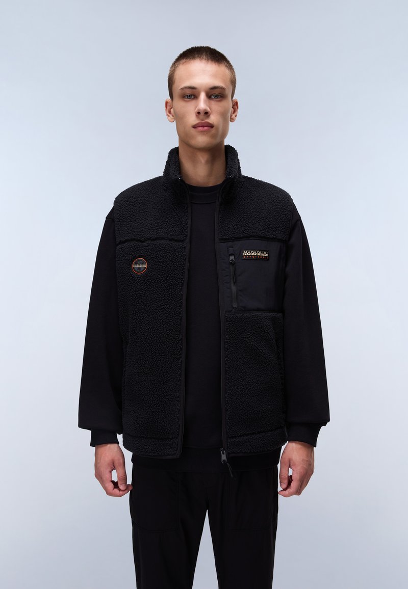 Black fleece vest with a high collar, textured surface, front zipper, and logo patch. Worn over a black long-sleeve shirt and black pants.