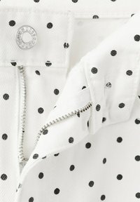 Tissu blanc à pois noirs avec une fermeture éclair argentée partiellement dézippée et une fermeture à bouton-pression argentée.