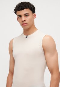 Haut sans manches beige en tissu texturé. Possède un col rond et une petite étiquette noire au centre devant. Design ajusté.