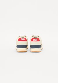 Beige sneakers med røde aksenter og merkevarelogo på baksiden, med en teksturert marinblå såle og glatte overflater.