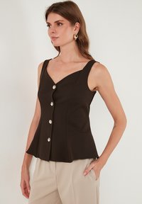 Rjava peplum bluza z globokim V-izrezom, ki ima velike srebrne gumbe spredaj in široke naramnice. Gladka tekstura tkanine.