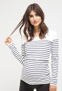 Femme aux cheveux bruns ondulés portant un haut blanc à manches longues avec des rayures horizontales bleu marine, tenant une mèche de ses cheveux, sur un fond uni.