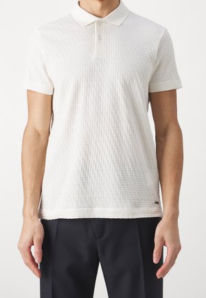 Man draagt een wit, gestructureerd, kortärmelig poloshirt gecombineerd met een zwarte broek, staand tegen een effen achtergrond.