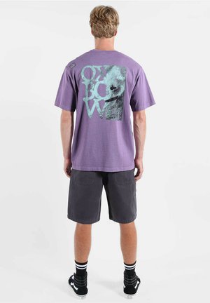 T-shirt en coton violet avec un grand motif graphique turquoise comprenant des lettres au dos, associé à un short gris et des baskets noires.