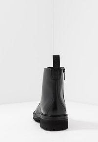 Bottine en cuir noir avec une finition lisse, un talon renforcé, une fermeture à glissière latérale et une tirette avec la marque "HUGO" à l'arrière.