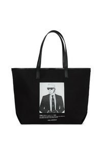 Crna torba na rame s dva duga rukohvata, koja prikazuje bijeli grafički print muškarca u sunčanim naočalama i odijelu. Izrađena od izdržljive tkanine.
