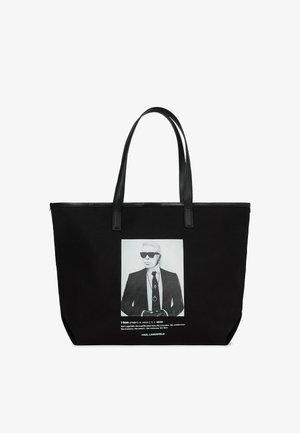 Crna torba na rame s dva duga rukohvata, koja prikazuje bijeli grafički print muškarca u sunčanim naočalama i odijelu. Izrađena od izdržljive tkanine.