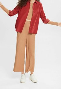 Chemise en velours côtelé couleur rouille portée sur un crop top côtelé marron clair et un pantalon large. Des baskets blanches complètent la tenue.