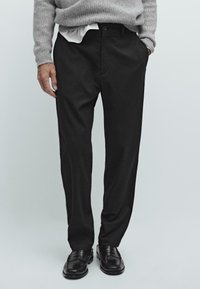 Man in zwarte formele broek, zwarte leren loafers, een grijze gebreide trui en een witte overhemd in de taille gestoken.