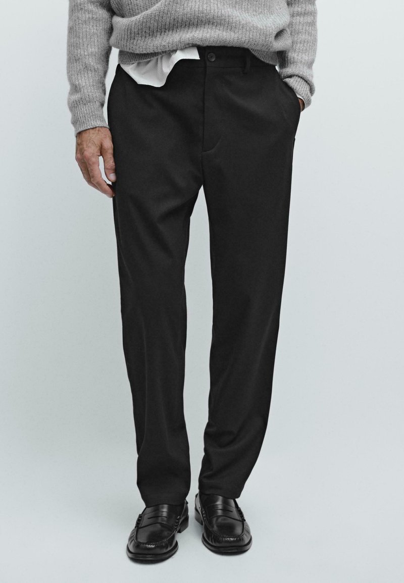 Man in zwarte formele broek, zwarte leren loafers, een grijze gebreide trui en een witte overhemd in de taille gestoken.