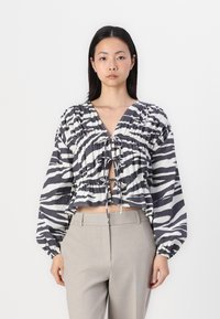 Blusa cropped riscada em preto e branco com um decote em V profundo, fecho com cordão e mangas volumosas, combinada com calças cinza claro ajustadas.