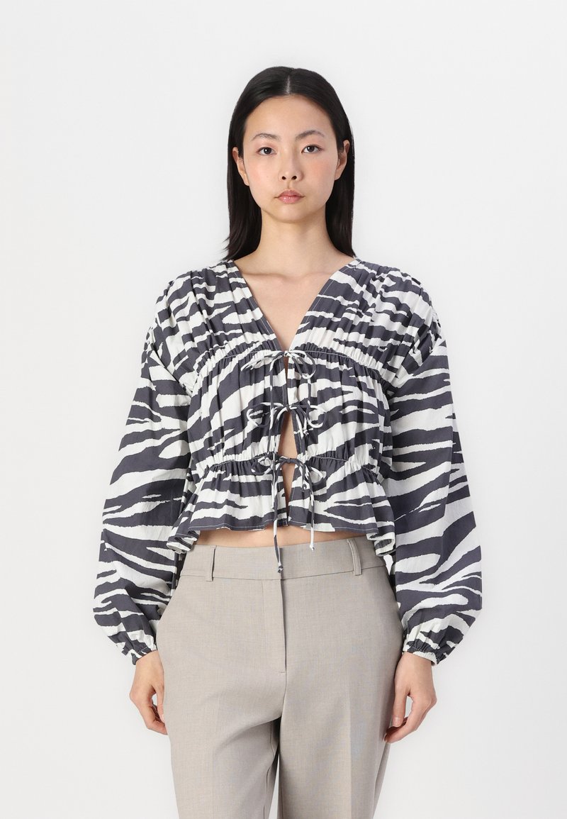 Blusa cropped riscada em preto e branco com um decote em V profundo, fecho com cordão e mangas volumosas, combinada com calças cinza claro ajustadas.