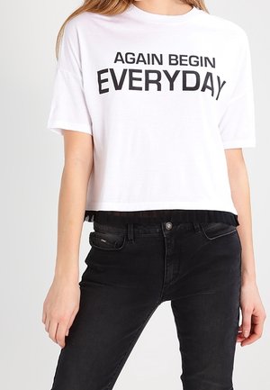 Witte cropped T-shirt met de tekst "AGAIN BEGIN EVERYDAY", gedragen met een zwarte jeans met hoge taille door een persoon die tegen een effen achtergrond staat.