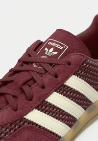 Μπορντό αθλητικό παπούτσι από σουέτ και δίχτυ με λευκό σχέδιο τριών ρίγων. Διαθέτει υφή επιφάνειας, κλείσιμο με κορδόνια και ετικέτα γλώσσας με λογότυπο.