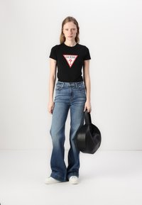 Black t-shirt with red triangular logo, distressed light blue flared jeans, and a round black handbag. Model stands against a plain background.