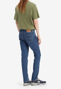 Personne portant une chemise manches courtes vert olive, un jean bleu en denim, et des baskets New Balance noires et blanches, debout de dos.
