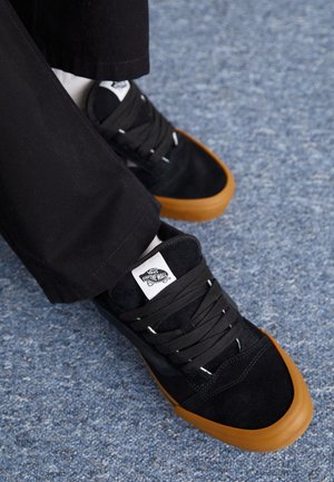 Zwarte suède sneakers met rubberen bruine zolen, voorzien van een wit logolabel en zwarte veters, gedeeltelijk bedekt door een paar zwarte broeken.