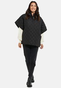 Svart vadderad poncho med krage och tryckknappar, bärs över en krämfärgad tröja. I kombination med svarta jeans och ankelstövlar.