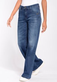 Jeans de mezclilla de pierna ancha en un azul oscuro, con cierre de botón, bolsillos frontales y un ligero desgaste. Llevados con unas zapatillas blancas para crear contraste.