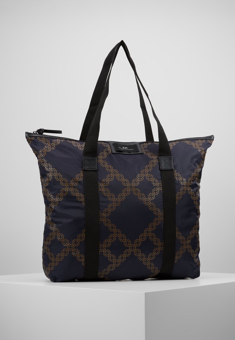 Sac fourre-tout bleu marine avec motif géométrique marron, bretelles noires et fermeture éclair, posé sur un piédestal blanc sur fond gris clair.