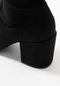 Stivaletto in suede nero con tacco spesso, caratterizzato da una texture liscia e un design senza cuciture. Il tacco è leggermente più largo alla base.
