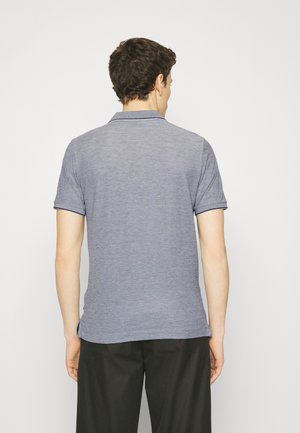 Homme de dos portant un polo gris à manches courtes avec bordures noires et un pantalon noir, sur un fond blanc uni.