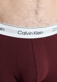 Burgundy férfi alsónemű fehér elasztikus derékkal, rajta a "Calvin Klein" logóval. Sima textúrájú és testhez simuló dizájn.