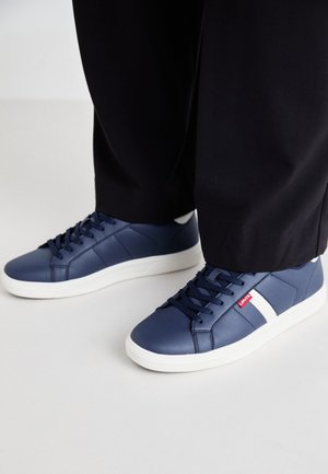 Zapatillas de deporte azul marino hechas de material sintético suave, con acentos blancos y una suela de goma texturizada. Cordones y marca visibles.