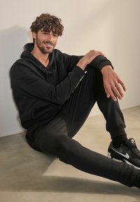 Svart hoodie och mjukisbyxor med elastiska manschetter. Modell som sitter mot en vägg, iklädd svarta sneakers med vita accenter. Slätt golvutseende.