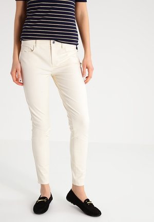 Personne portant un pantalon slim crème, un haut à manches courtes rayé bleu marine et blanc, et des mocassins noirs sans lacets, debout sur un sol blanc.