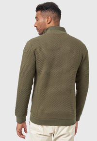 Suéter verde oliva hecho de un tejido texturizado con un patrón elevado, que presenta un cuello alto y puños y dobladillo acanalados.