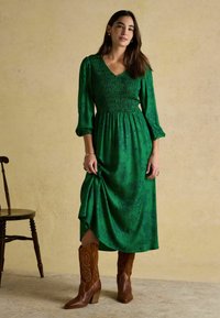 Žena v zelených midi šatách s dlhým rukávom a kvetinovým vzorom, s hnedými koženými kovbojskými čižmami, stojí pri drevenej stoličke.