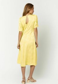 Gelbes Kleid mit kurzen Puffärmeln, floralem Muster, einem Schlüsselrücken und einem knielangen Saum. Getragen mit beigen Blockabsatzsandalen.