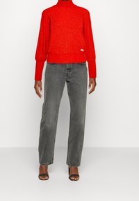 Vrouw die een felrode coltrui draagt, grijze rechte jeans en zwarte open-toe hoge hakken tegen een effen witte achtergrond.