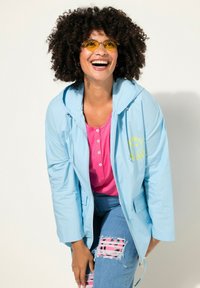 Chaqueta cortavientos azul claro con capucha y gráfico de carita sonriente amarilla, llevada sobre una camiseta rosa. Jeans de mezclilla desgastados con detalles de parches coloridos.