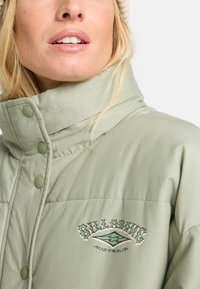 Nahaufnahme einer Person, die eine hellgrüne Jacke mit hohem Kragen und einem Billabong Australien-Logo auf der Brust trägt.