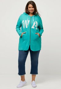 Sudadera de color verde azulado con cremallera y un gráfico blanco, bolsillos frontales y capucha con cordón, combinada con jeans cortos azul oscuro y zapatillas blancas.