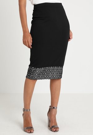 Vrouw die een zwarte kokerrok tot op de knie draagt met een patroon aan de zoom en zilveren hooggehakte sandalen met enkelbandje, staand op een witte vloer.