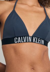 Bra triangolare grigio scuro con spalline sottili e un elastico nero con la scritta "CALVIN KLEIN" in lettere bianche. Texture liscia, design minimale.