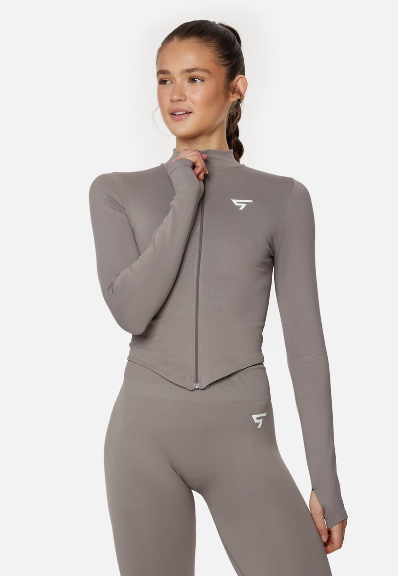 Light grey athletic long-sleeve top with a zip front, high collar, and thumbholes. Features a white logo on the chest. Smooth texture.
