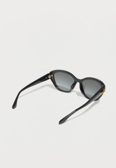Lunettes de soleil noires forme œil de chat avec verres teintés foncés et accents dorés sur les branches, posées sur une surface blanche.