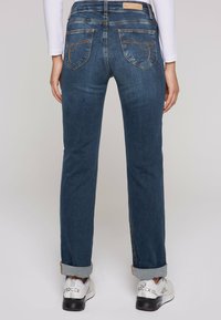 Jeans de mezclilla azul oscuro con pierna recta, puños doblados y costuras decorativas en los bolsillos traseros. Zapatillas con detalles plateados.