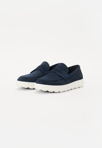 Des chaussures slip-on bleu marine en daim, avec une épaisse semelle blanche, un design incurvé et un détail de sangle texturé sur le dessus.