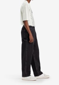 Hombre vistiendo jeans negros holgados, camiseta gráfica blanca y zapatillas blancas, de perfil lateral contra un fondo blanco plano.