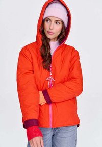 Kobieta w jasnopomarańczowej kurtce z kapturem, założonej na różowy sweter i czapkę, z warstwowymi czerwonymi i pomarańczowymi rękawami, stoi z założonymi rękami.