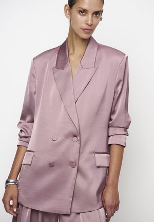 Vrouw die een oversized zijden lavendelkleurige double-breasted blazer draagt met opgerolde mouwen en een bijpassende broek, staand tegen een witte achtergrond.