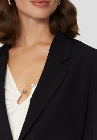Zwarte blazer met een getailleerd ontwerp, voorzien van een gekarteld kraag en gestructureerde schouders, gecombineerd met een witte top, accentueert met een pareldetail.
