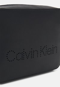 Borsa nera in pelle con una texture liscia e un logo traforato con la scritta "Calvin Klein" sul davanti. Forma rettangolare.